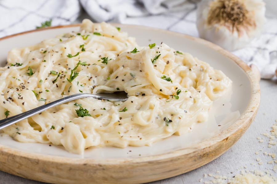 A white and bamboo plate filled with fettucine alfredo nearby ground parmesan and a garlic bulb.