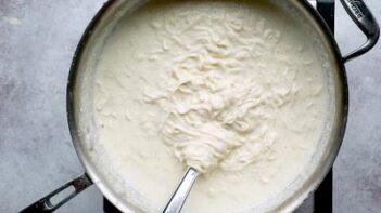 A whisk stirring noodles into a skillet of creamy white alfredo sauce.