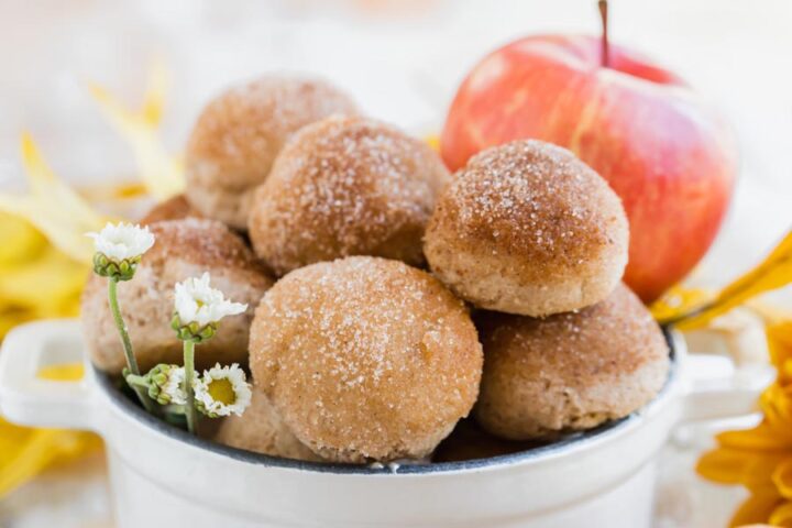 A white dish filled with keto apple cider donut bites, topped with cinnamon “sugar” and garnished with small white flowers and a red apple in the background.
