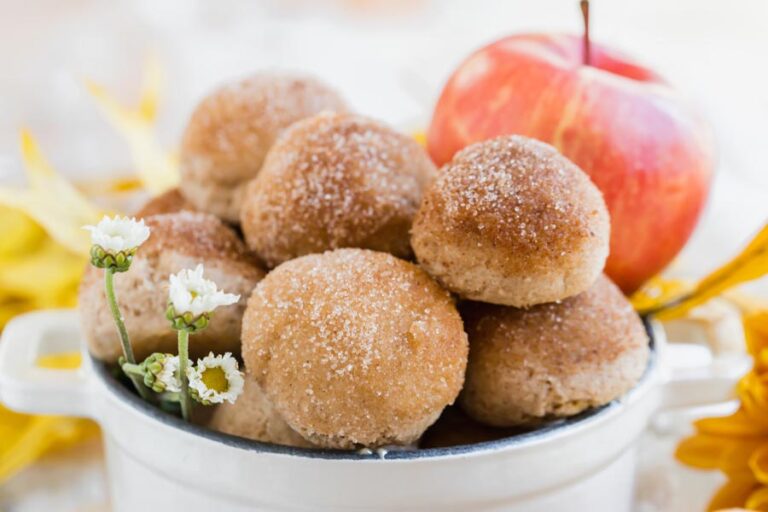 A white dish filled with keto apple cider donut bites, topped with cinnamon “sugar” and garnished with small white flowers and a red apple in the background.