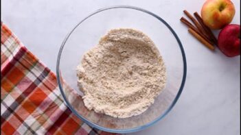 Overhead shot of a glass bowl filled with almond flour mixture on a white surface next to apples, cinnamon sticks, and a plaid towel.