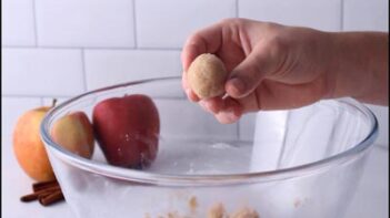 Close-up of a hand holding a formed dough ball over an empty glass bowl with apples in the background.