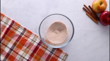 Bowl of cinnamon “sugar” coating next to apples and cinnamon sticks on a plaid cloth.