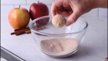 Hand dipping a baked donut bite into a bowl of cinnamon “sugar” coating.