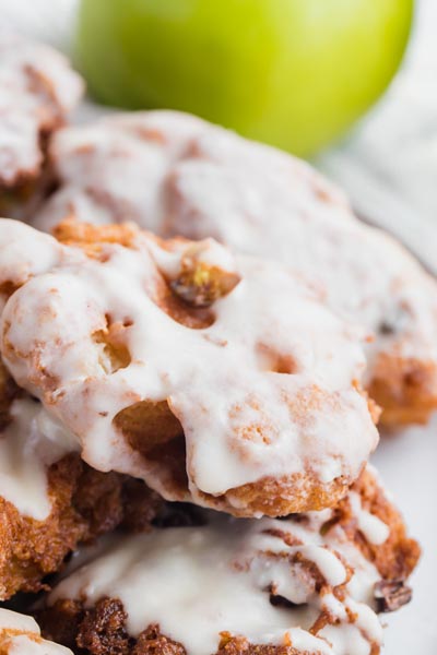 An glazed apple fritter on top of other fritters on a plate with a green apple in the background.