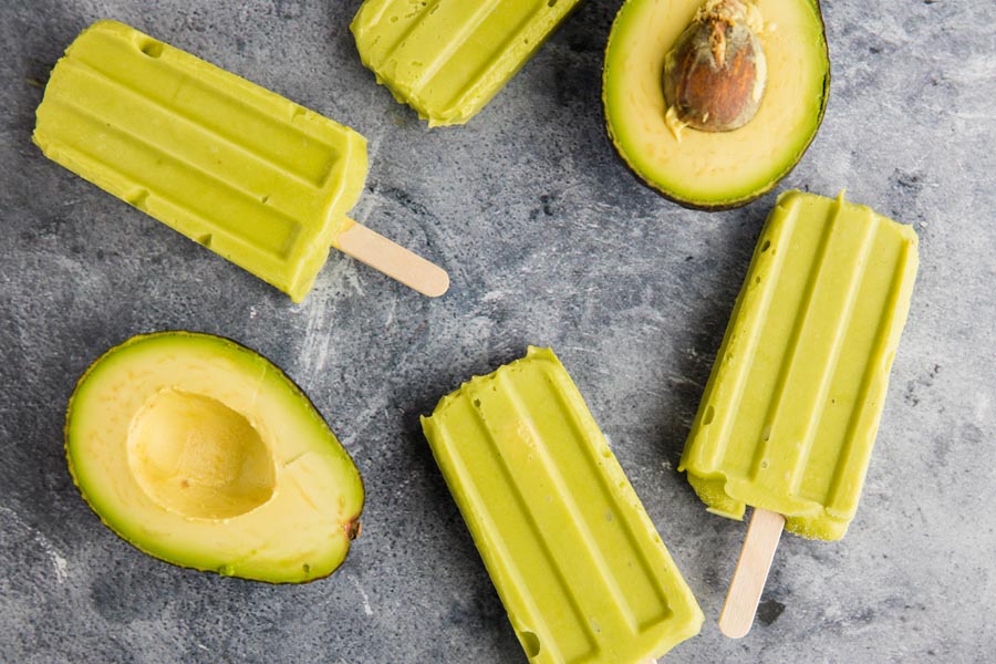 creamy avocado popsicles on a black background with halved avocados scattered around