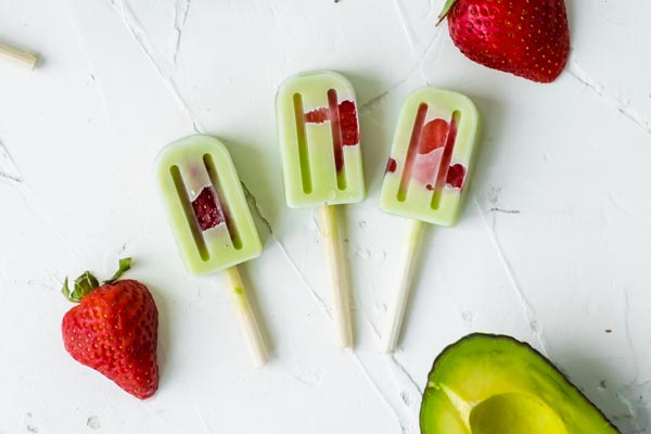 keto strawberry avocado popsicles on a white background with fresh strawberries and slice avocado nearby