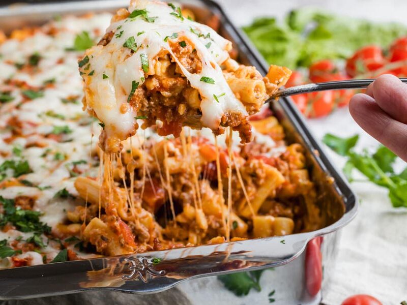 A hand pulling out a serving of pasta casserole from the dish.
