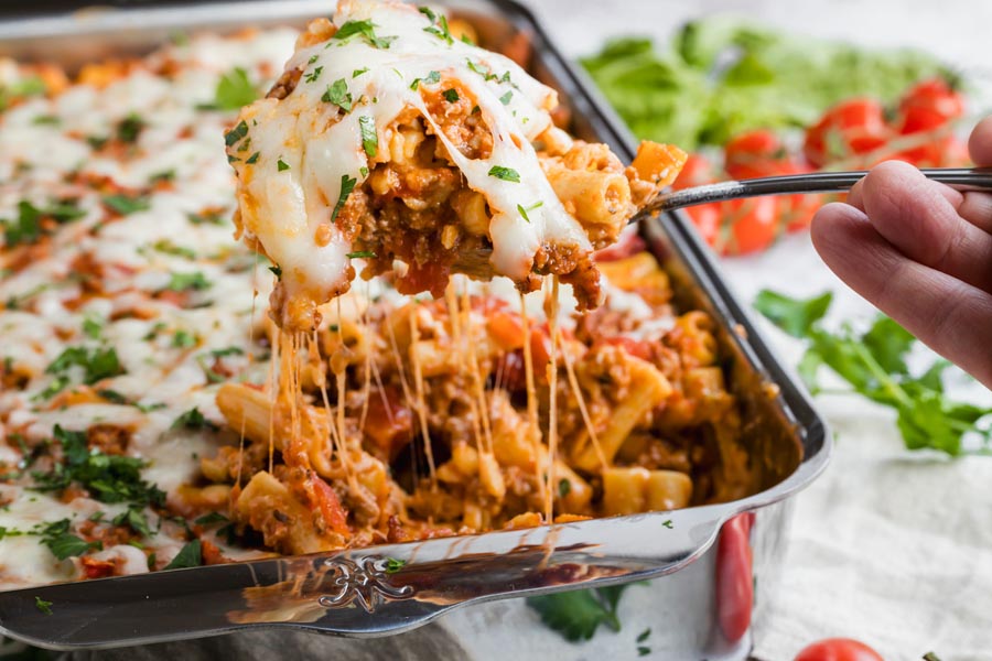 A hand pulling out a serving of pasta casserole from the dish.