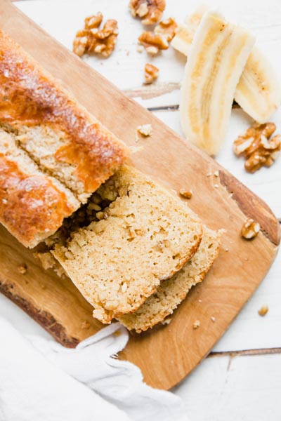 two slices of banana bread sliced on a wooden cutting board with walnuts and bananas above