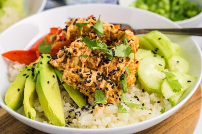 Crispy bang bang chicken on top of cauliflower rice next to avocado, cucumber and topped with sauce and sesame seeds.