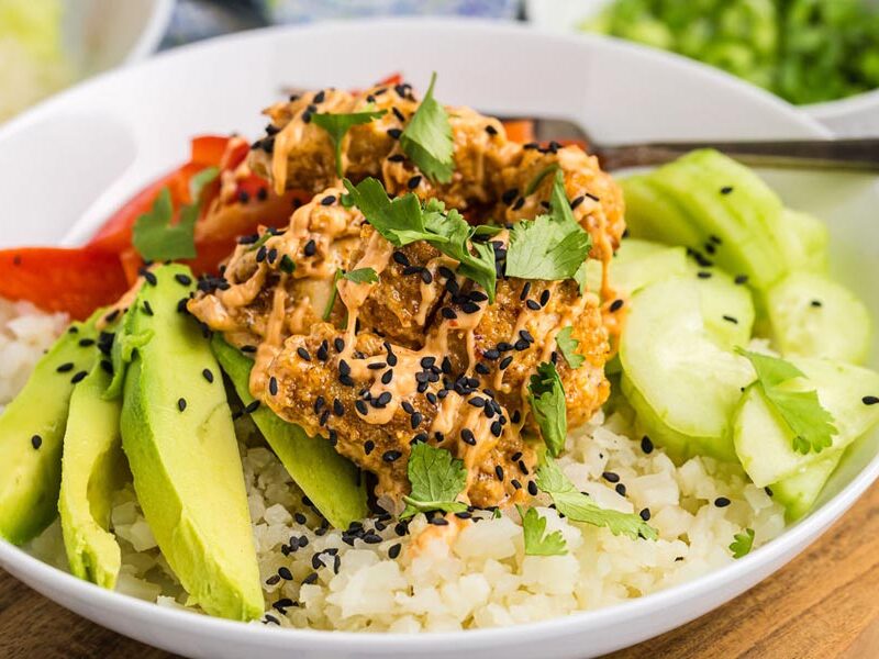 Crispy bang bang chicken on top of cauliflower rice next to avocado, cucumber and topped with sauce and sesame seeds.