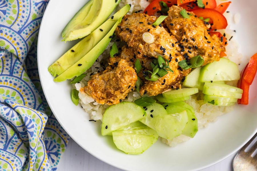 Fried chicken covered in bang bang sauce and served on a plate next to cucumber, red pepper and avocado.