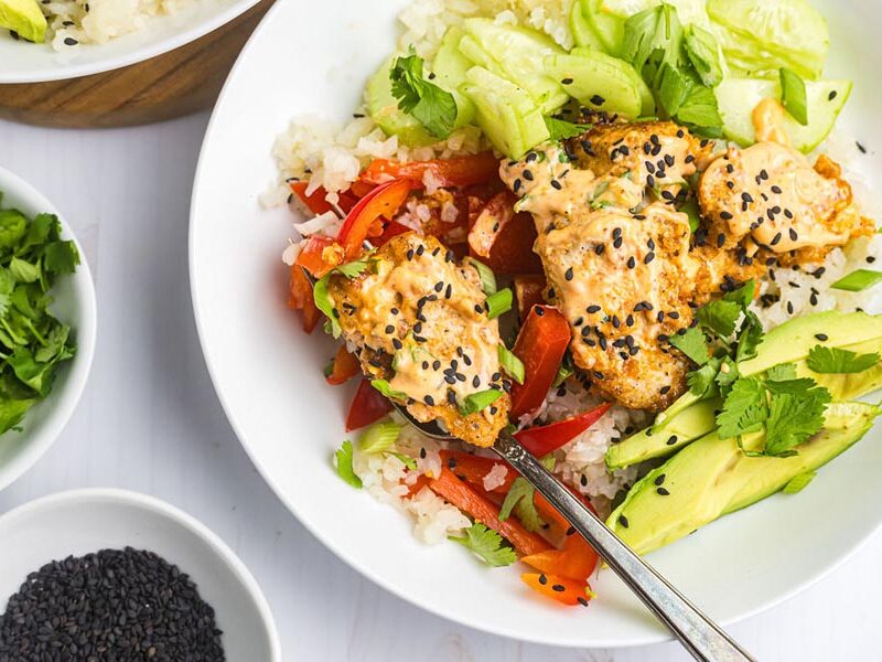 A fork holding a piece of bang bang chicken over a bowl with cauli rice and veggies.