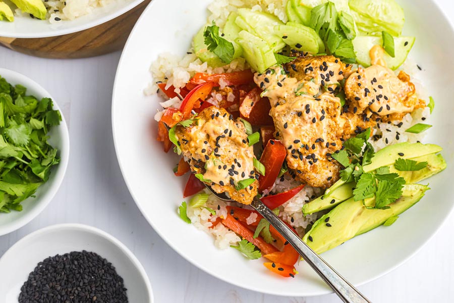 A fork holding a piece of bang bang chicken over a bowl with cauli rice and veggies.