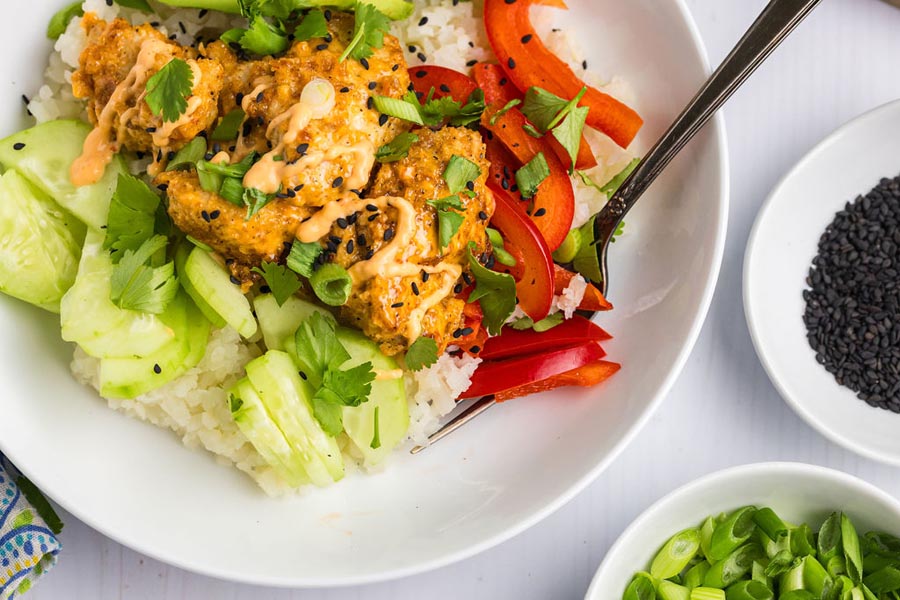 Bang bang chicken bowl on a plate next to sesame seeds and green onions.