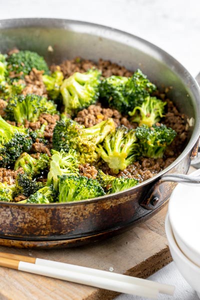 a large skillet with crispy beef and broccoli florets next to chopsticks