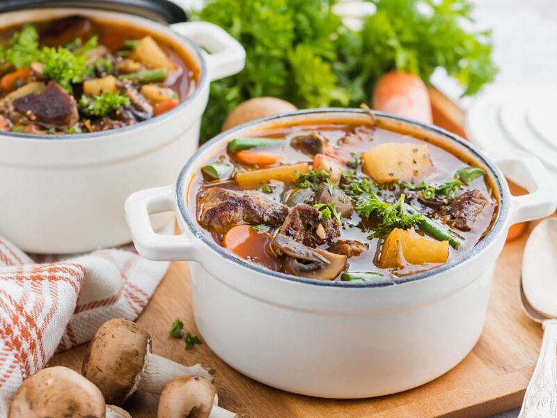 Two white pots of beef stew with carrots, mushrooms, green beans and parsley.