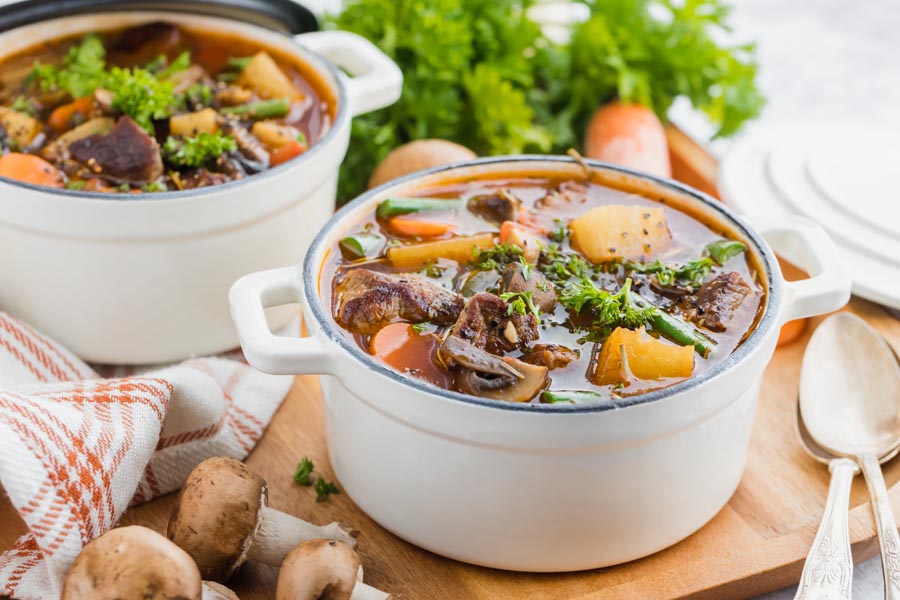 Two small dutch oven pots filled with beef stew with two spoons to the right and fresh whole tomatoes to the left.