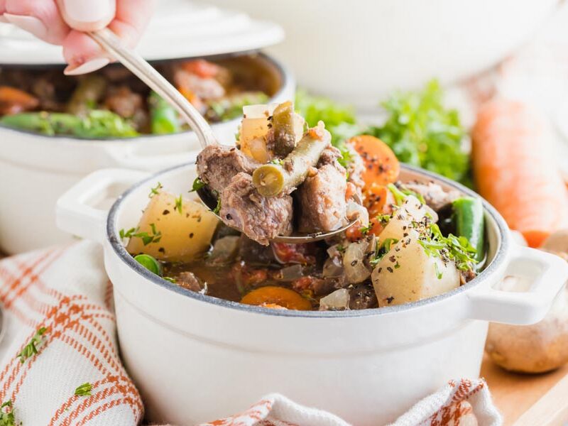 a hand holding a spoonful of beef stew with chunks of beef and green beans in it.