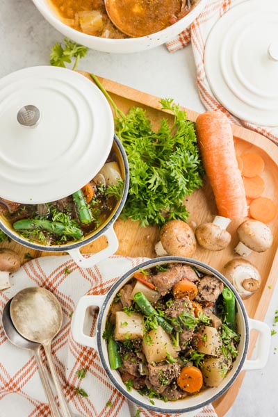 Top down view of a small bowl of stew filled with beef, carrot, green beans and daikon radish next to another pot with a lid on it. Various diced vegetables are nearby.