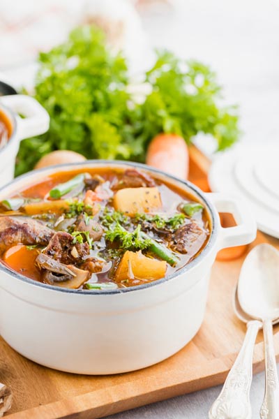 A pot of beef stew on a wooden board with carrots in the background and spoons nearby.
