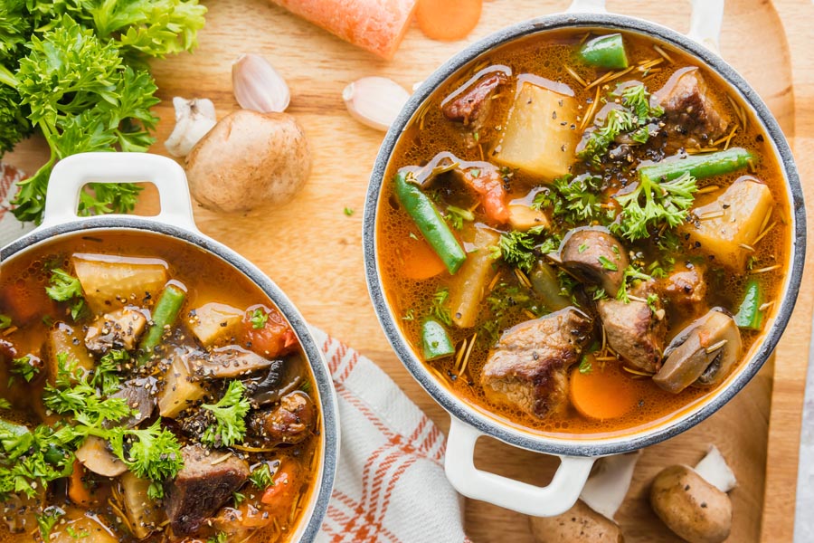 Looking down into two pots filled with chunky beef soup with lots of vegetables and chopped vegetables near by.