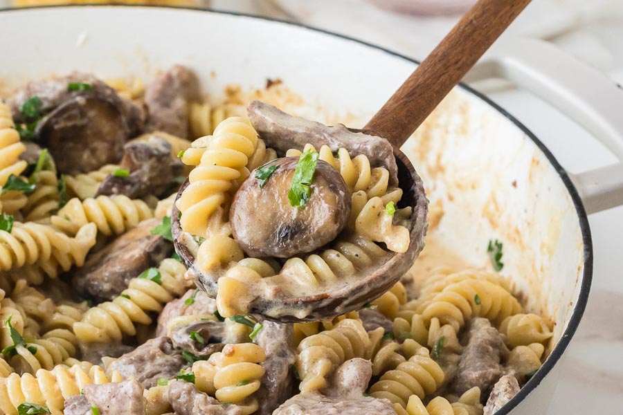 A wooden spoon holding creamy keto beef stroganoff with mushrooms, steak, and rotini pasta in a skillet.
