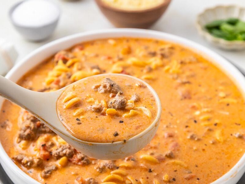 A ladle holding a scoop of tomato soup filled with noodles, ground beef and tomatoes.