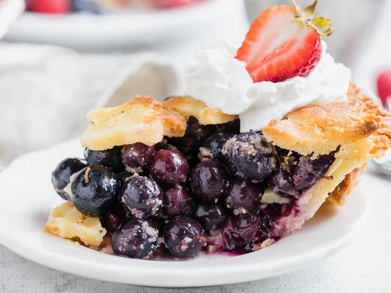 a slice of blueberry pie on a white plate topped with whipped cream and a strawberry
