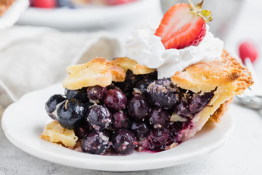 a slice of blueberry pie on a white plate topped with whipped cream and a strawberry