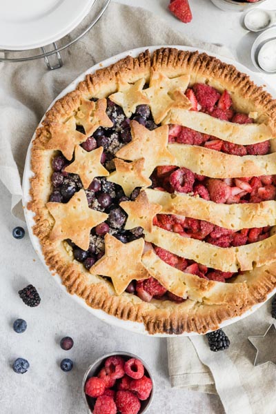A colorful red and blue berry pie with berries and measuring spoons nearby