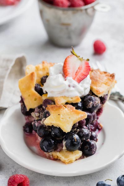 a slice of blueberry pie with stars on the top pie crust and topped with a dollop of whipped cream and a strawberry