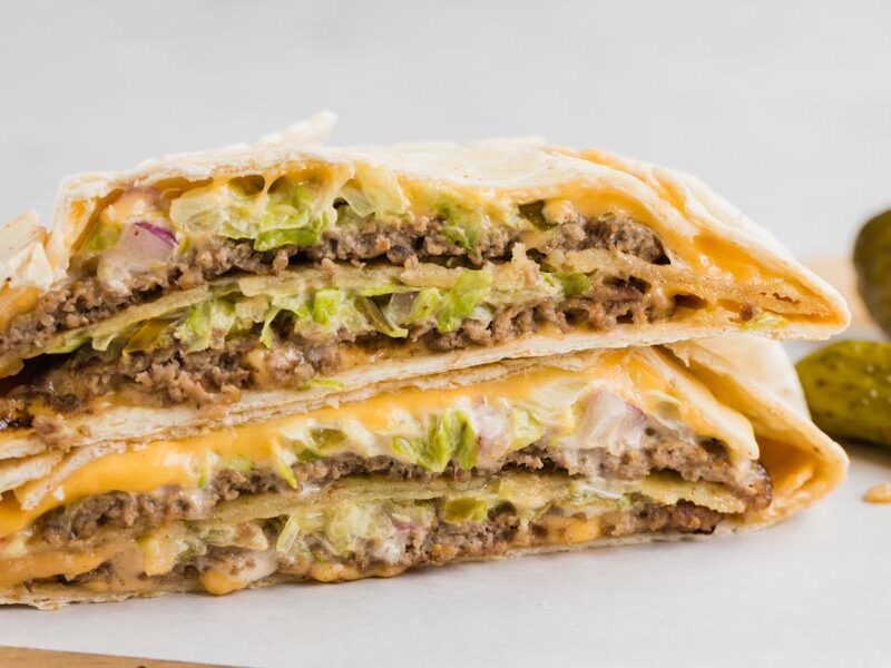 A big mac crunchwrap cut in half and stacked on each other with two dill pickles off to the side.