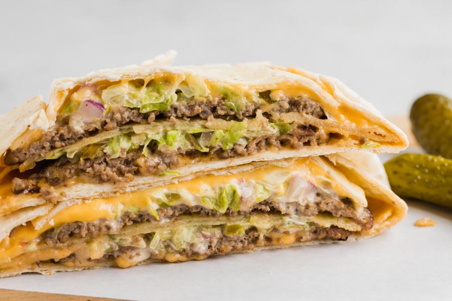 A big mac crunchwrap cut in half and stacked on each other with two dill pickles off to the side.