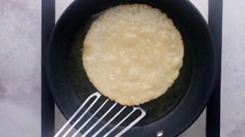 A tostada shell frying in a skillet while a spatula starts to lift it up.