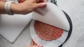 Peeling back a parchment circle to reveal a flat, thin beef patty that was flattened by a tortilla press.