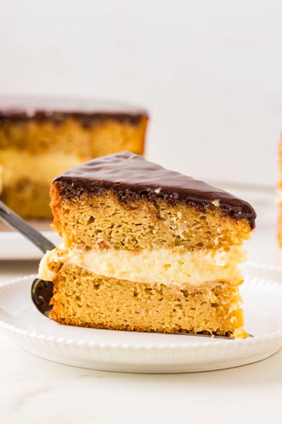 Single slice of keto Boston cream pie on a dessert plate, showcasing tender cake layers, vanilla custard filling, and chocolate ganache glaze.