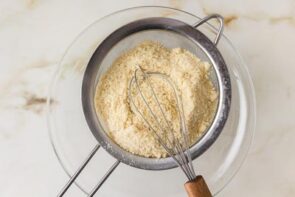 Fine powder and a whisk in a mesh strainer over a glass bowl.