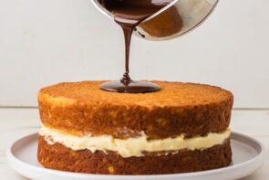 Chocolate ganache being poured over a layered keto Boston cream pie filled with vanilla custard.