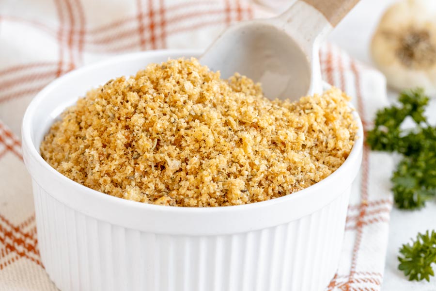 Keto panko breadcrumbs with Italian seasonings sits in a white bowl with fresh parsley and garlic nearby.