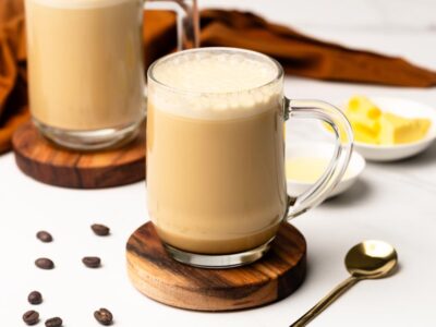 Creamy keto bulletproof coffee served in a glass mug with a frothy top, surrounded by coffee beans, butter, and cream.