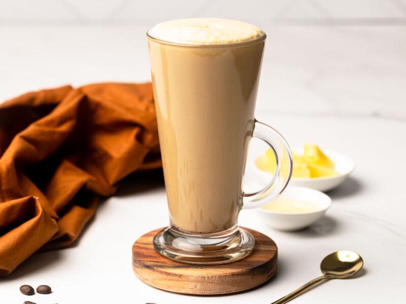 Tall glass of frothy keto bulletproof coffee served on a wooden coaster with butter and cream in the background.