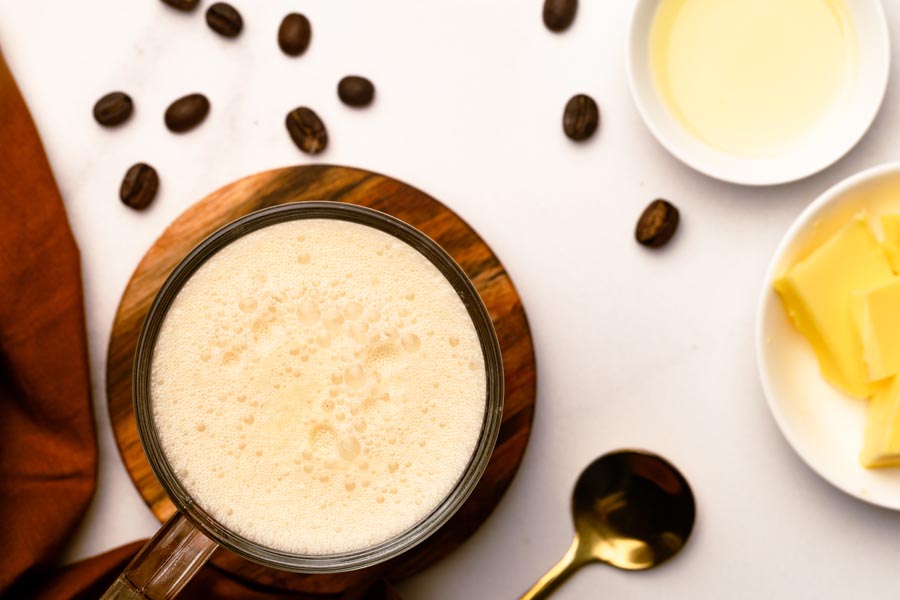 Tall glass of frothy keto bulletproof coffee served on a wooden coaster with butter and cream in the background.