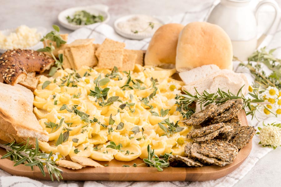 a beautiful butter board surrounded by various breads and crackers and mixed herbs