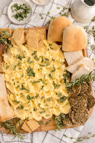 a party board of compound butter, sliced bread, rolls and crackers on a checkered towel next to fresh herbs