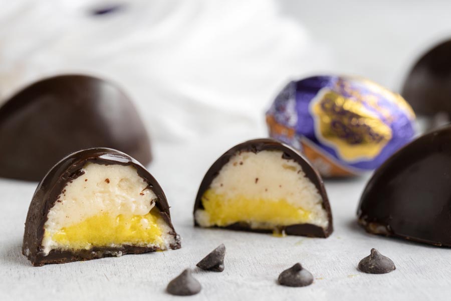 a creme egg sliced in half showing the gooey center with a whole cadbury foil wrapped egg in the background