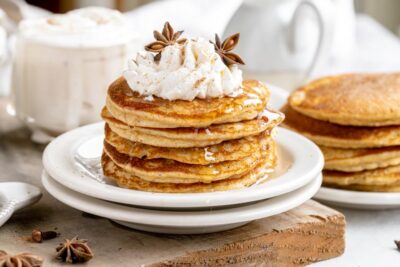 a stack of five pancakes on two white plates, topped with whipped cream and star anise with more pancakes behind