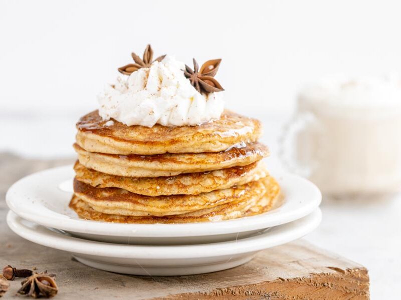 a stack of pancakes topped with whipped cream and spices with a chai latte in the background