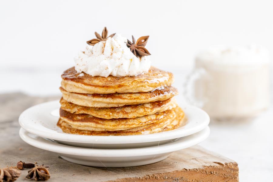 a stack of pancakes topped with whipped cream and spices with a chai latte in the background